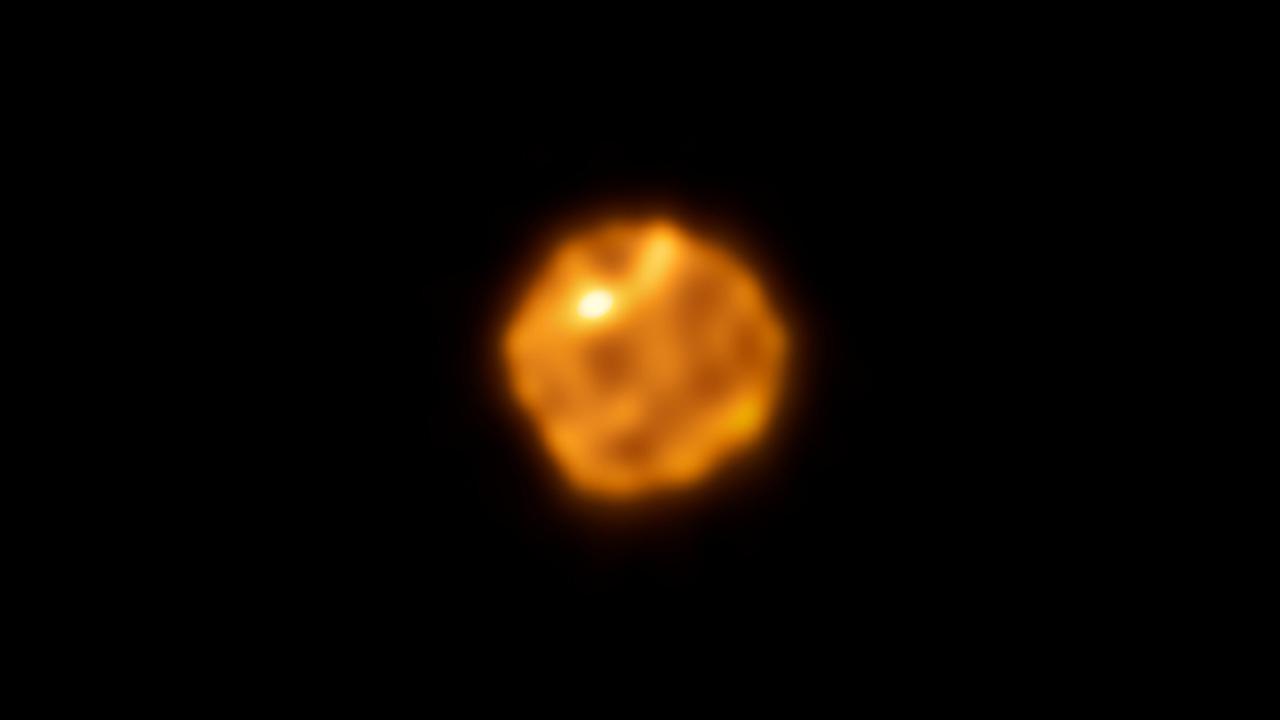Imagen de ALMA de la superficie submilimétrica de Betelgeuse: un disco granulado naranja y rojo, de contorno irregular, con una mancha brillante hacia el noreste.