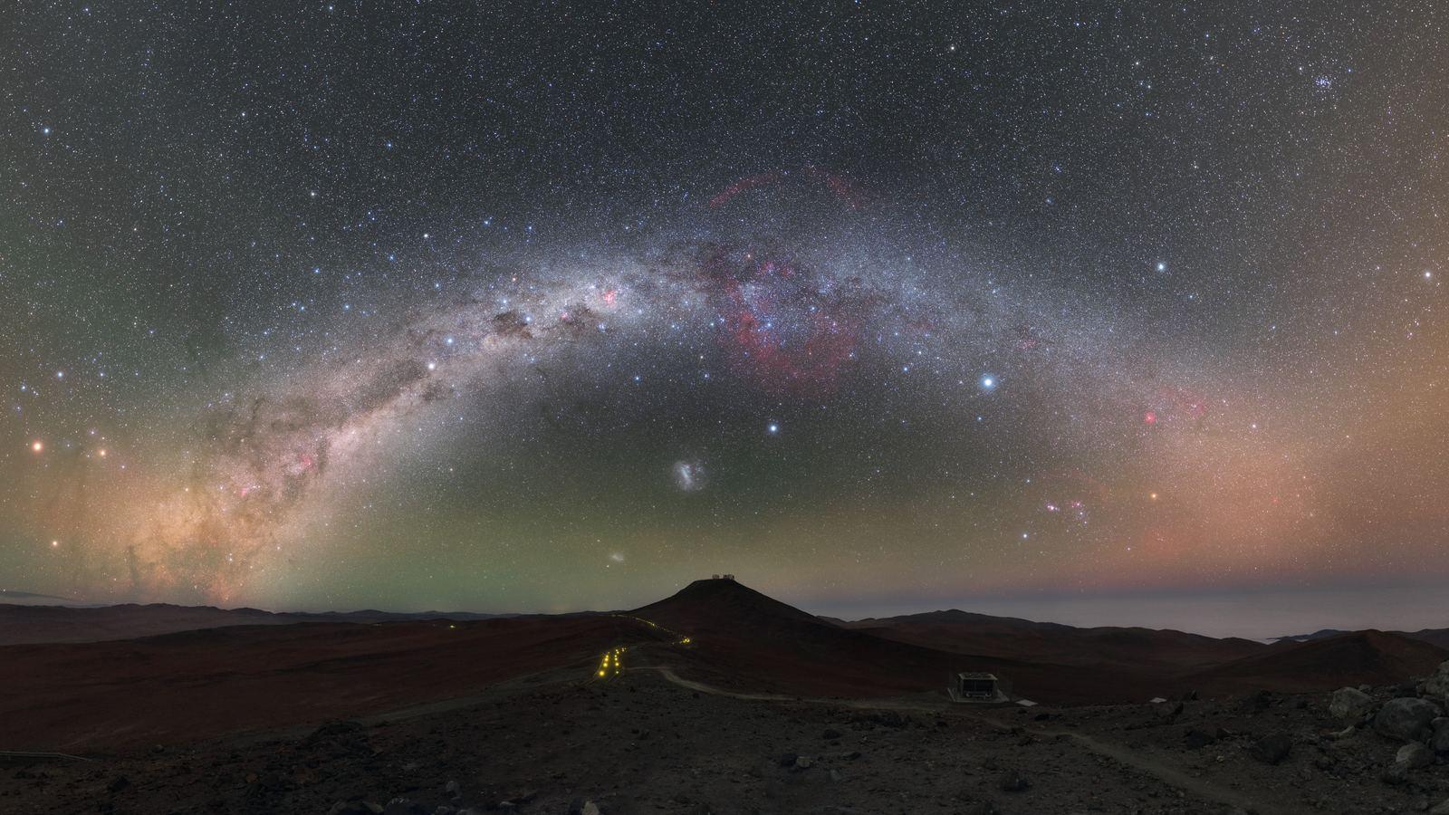 El cielo nocturno sobre el observatorio de Paranal, en Chile: la Vía Láctea en arco, tonos verdes y rojos del resplandor atmosférico y las cúpulas de los telescopios apenas iluminadas.