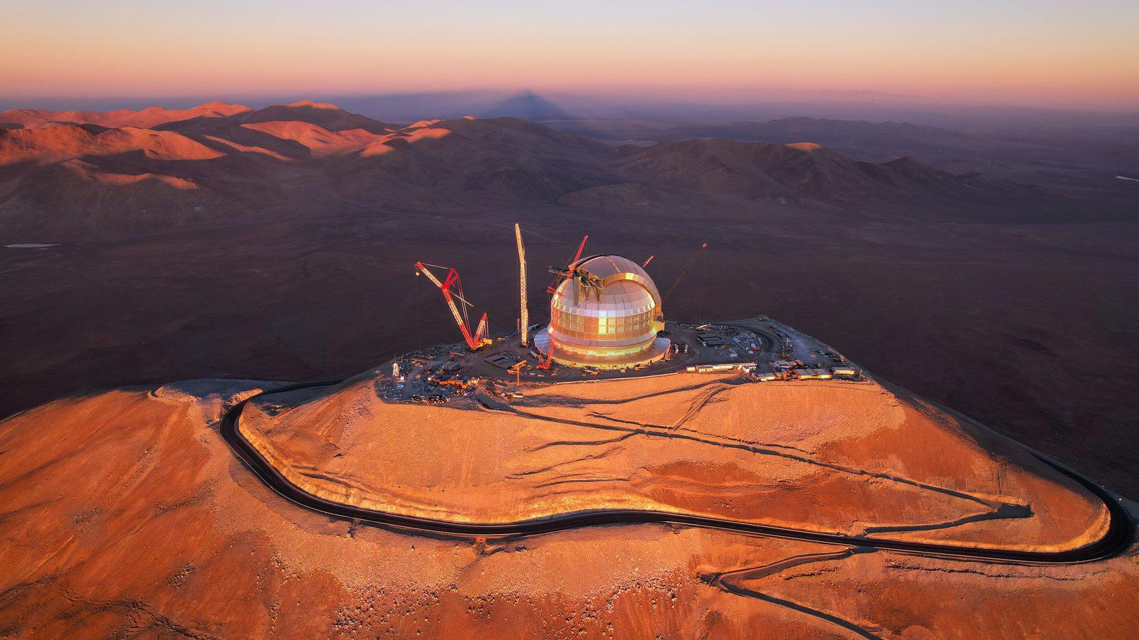 La cúpula en construcción del Extremely Large Telescope, iluminada por el sol poniente en el desierto de Atacama, rodeada de grandes grúas, con la montaña proyectando su sombra a lo lejos.