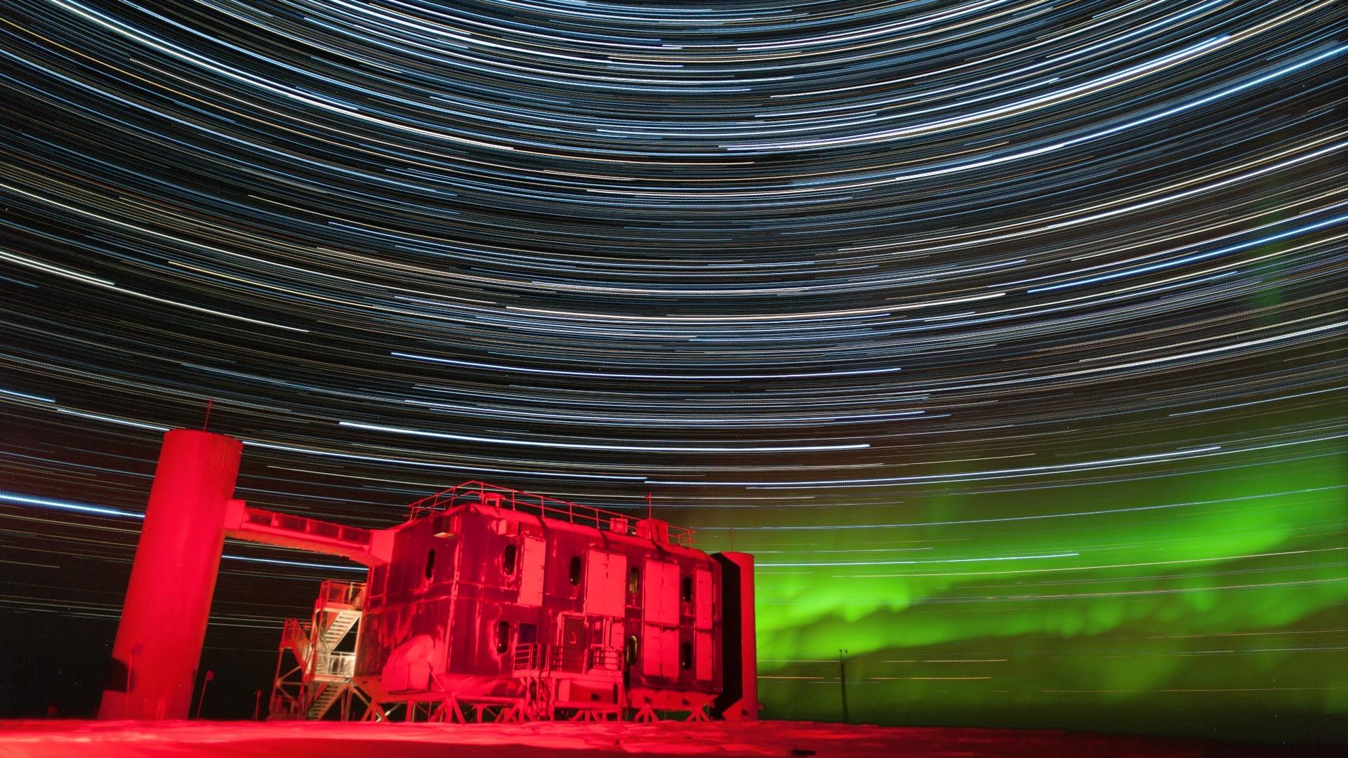 El laboratorio IceCube sobre el hielo del polo Sur, bajo un cielo nocturno cruzado por estelas circulares de estrellas y bañado por un resplandor verde.