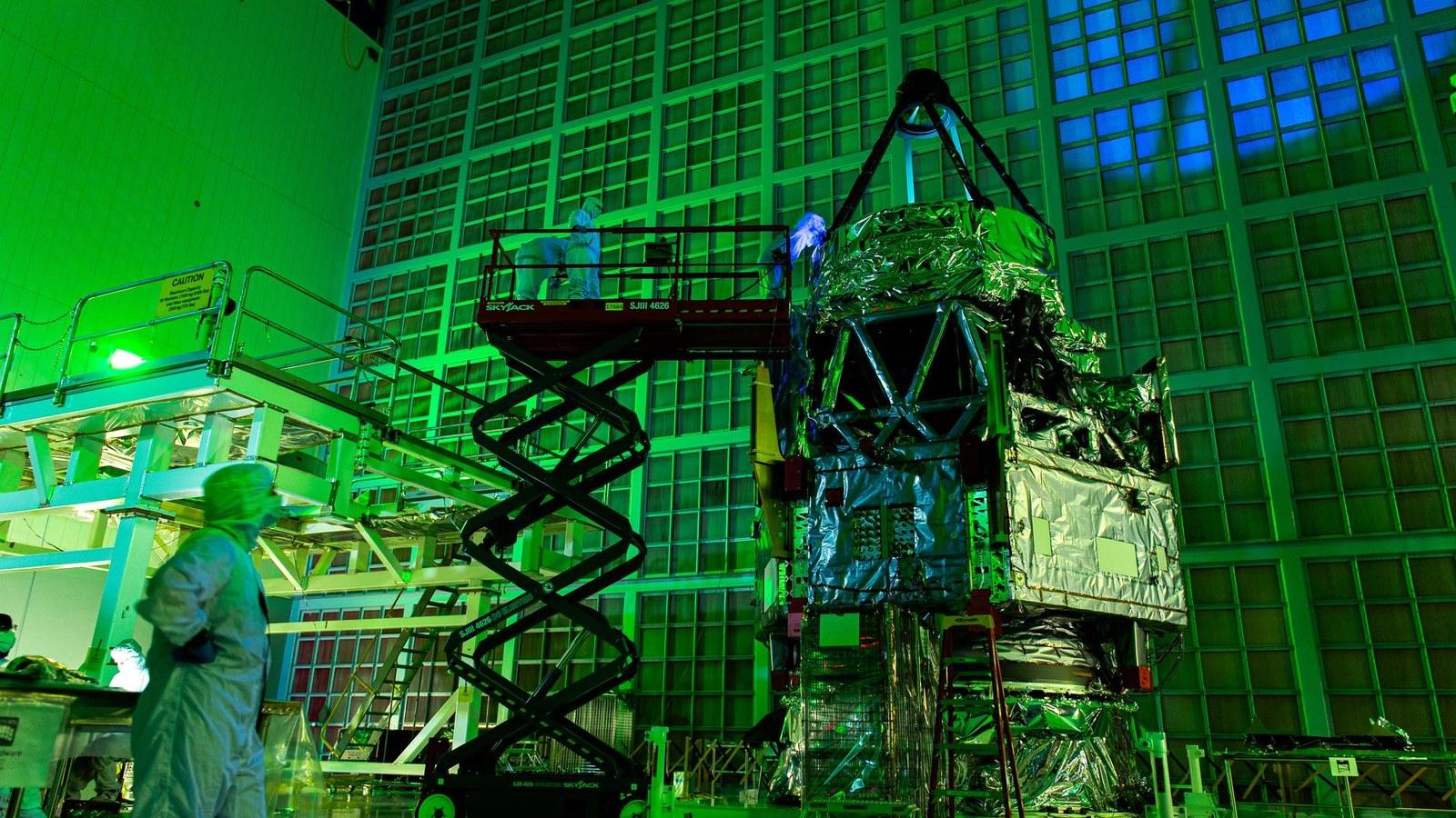 Roman team members on a scissor lift shine an ultraviolet light onto the telescope's primary mirror inside a clean room bathed in a green glow.