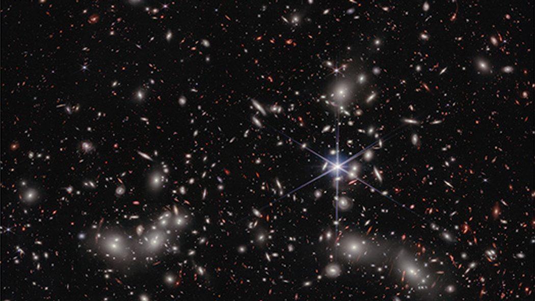 The galaxy UHZ1 imaged in infrared by the James Webb Space Telescope, a small orange galaxy adrift in a field of distant galaxies.