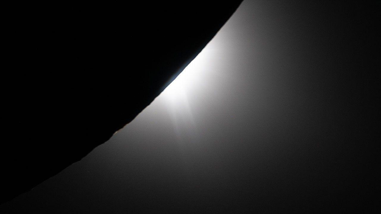The Moon in silhouette, dark, in front of an almost fully hidden solar disc: a thin thread of light outlines its edge, and a jet of plasma streams out of the solar corona.