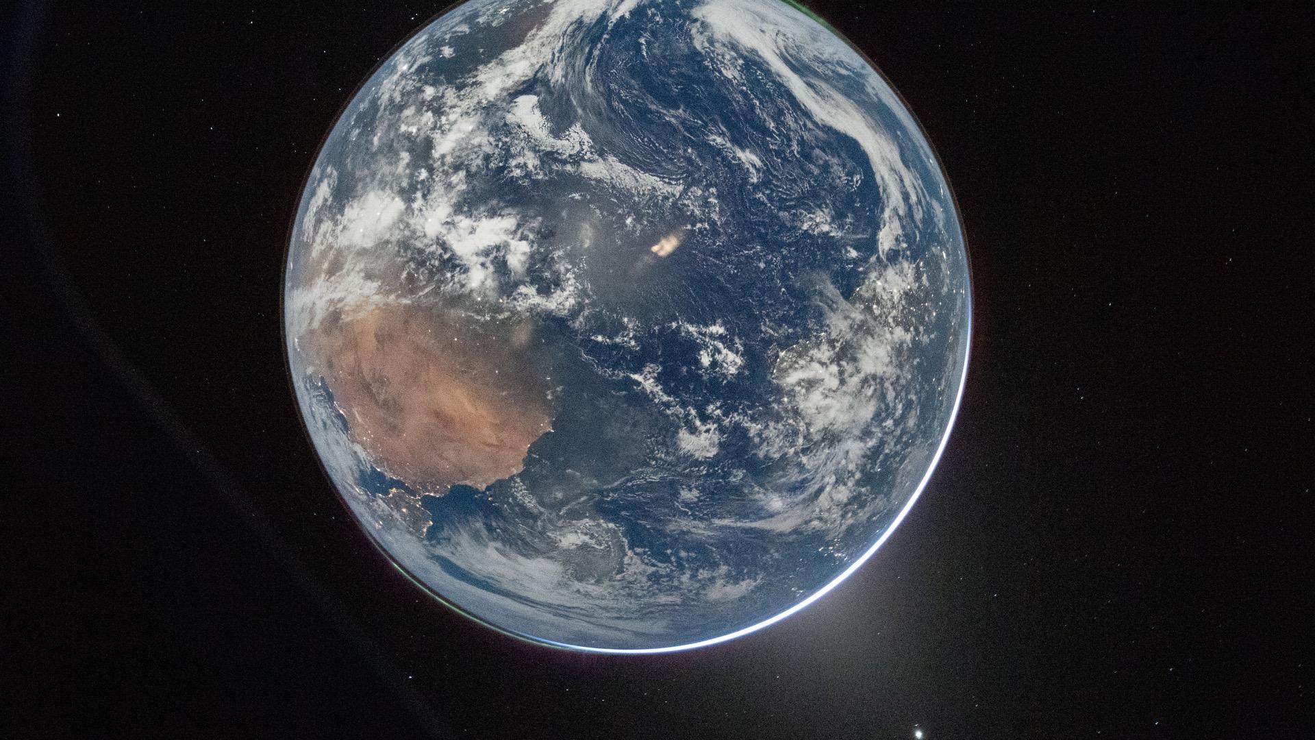 Earth at night photographed from space: a dark blue disc with shadowy continents and a thin film of atmosphere, the left half sunk in darkness with a few city lights.