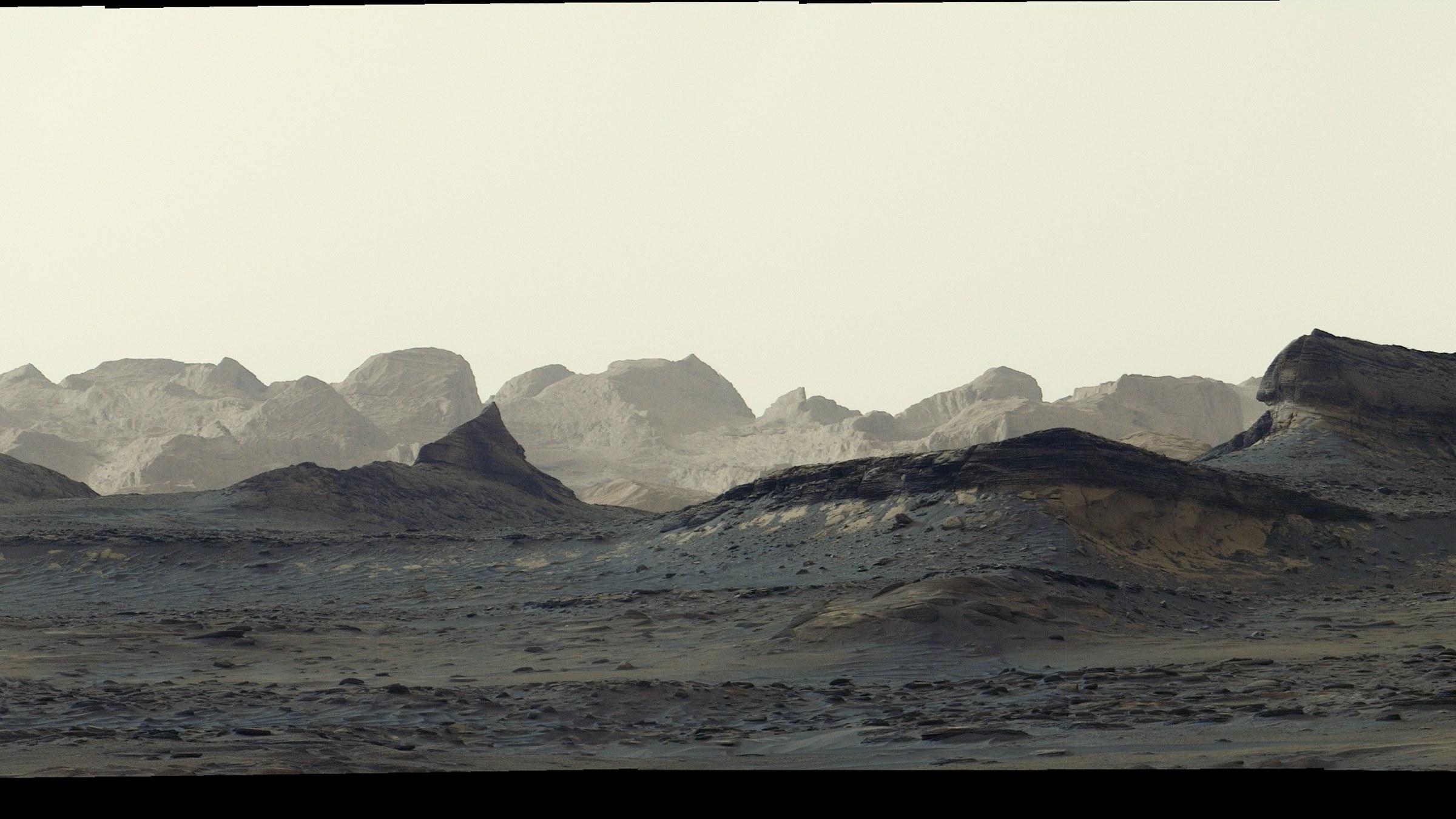Panorama de seis imágenes unidas por la NASA: al amanecer, unas crestas rocosas esculpidas por el viento se alzan en el horizonte del Monte Sharp, bajo un cielo y un suelo teñidos de azul.