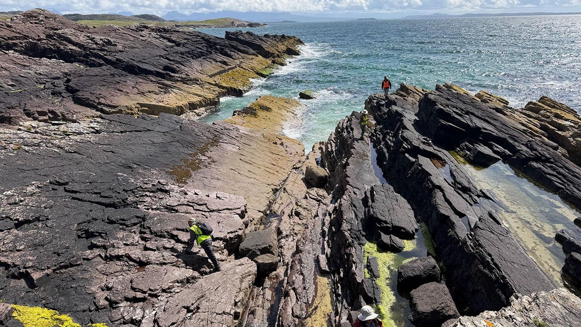 Investigadores con equipo de campo y mochilas al borde de un acantilado de arenisca roja sobre el océano, en Clachtoll, Escocia, bajo un cielo bajo y gris.