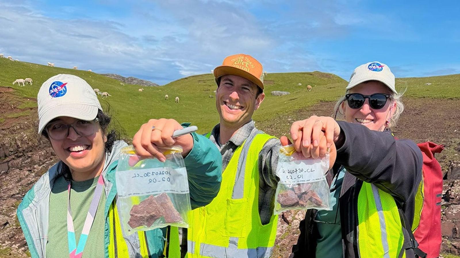 Tres miembros del equipo, con ropa de campo, sostienen uno al lado del otro fragmentos de roca roja llenos de manchas verdes, a la orilla del agua en Clachtoll Bay.