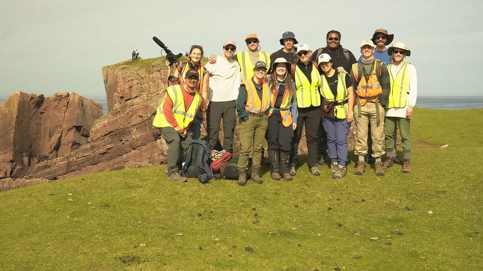 Foto de grupo de una docena de investigadores con ropa de campo, alineados en dos filas ante un paisaje rocoso escocés.