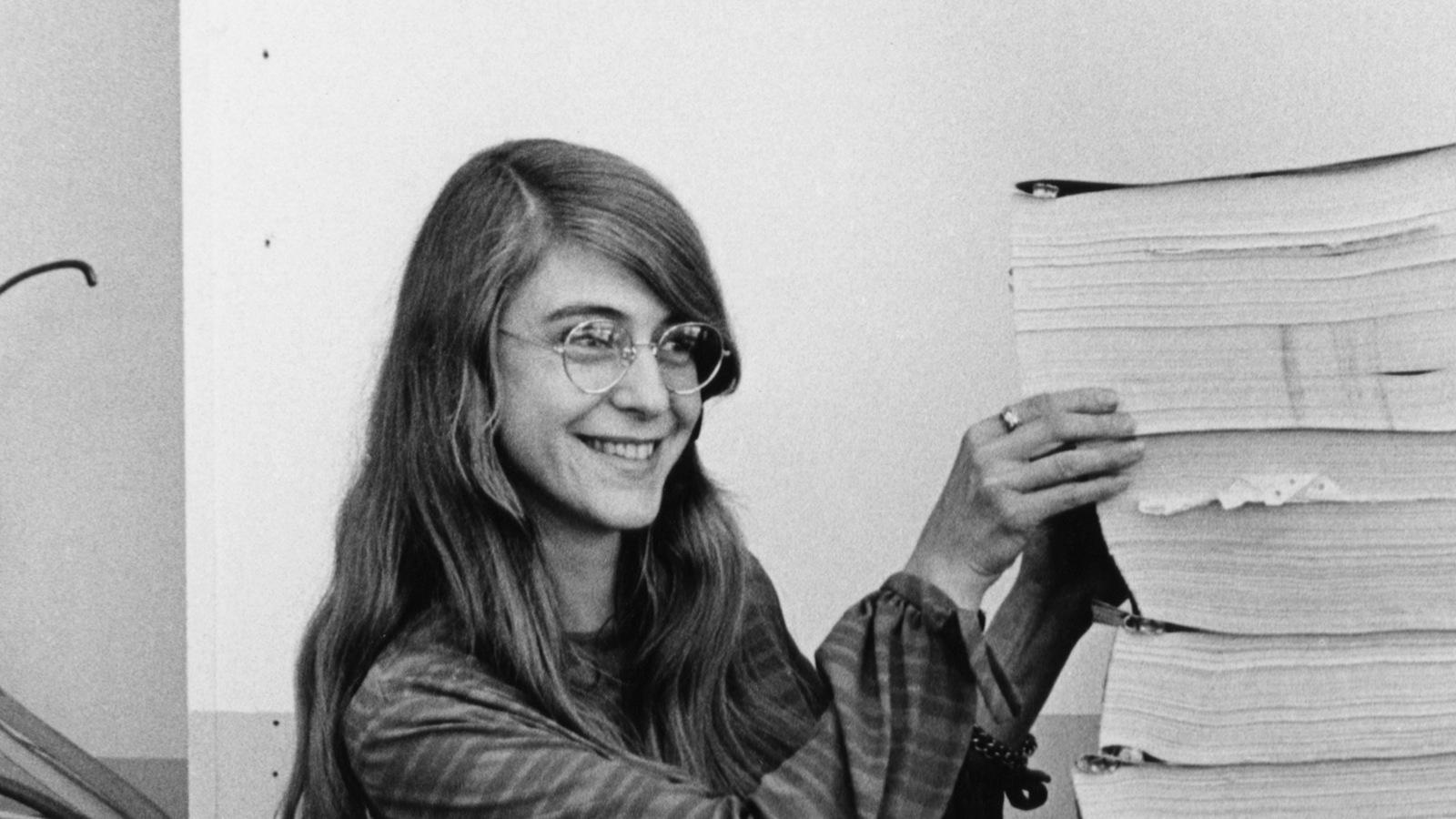 Margaret Hamilton standing in 1969 beside stacks of printed computer listings, the documentation for the Apollo flight software built by her MIT team.