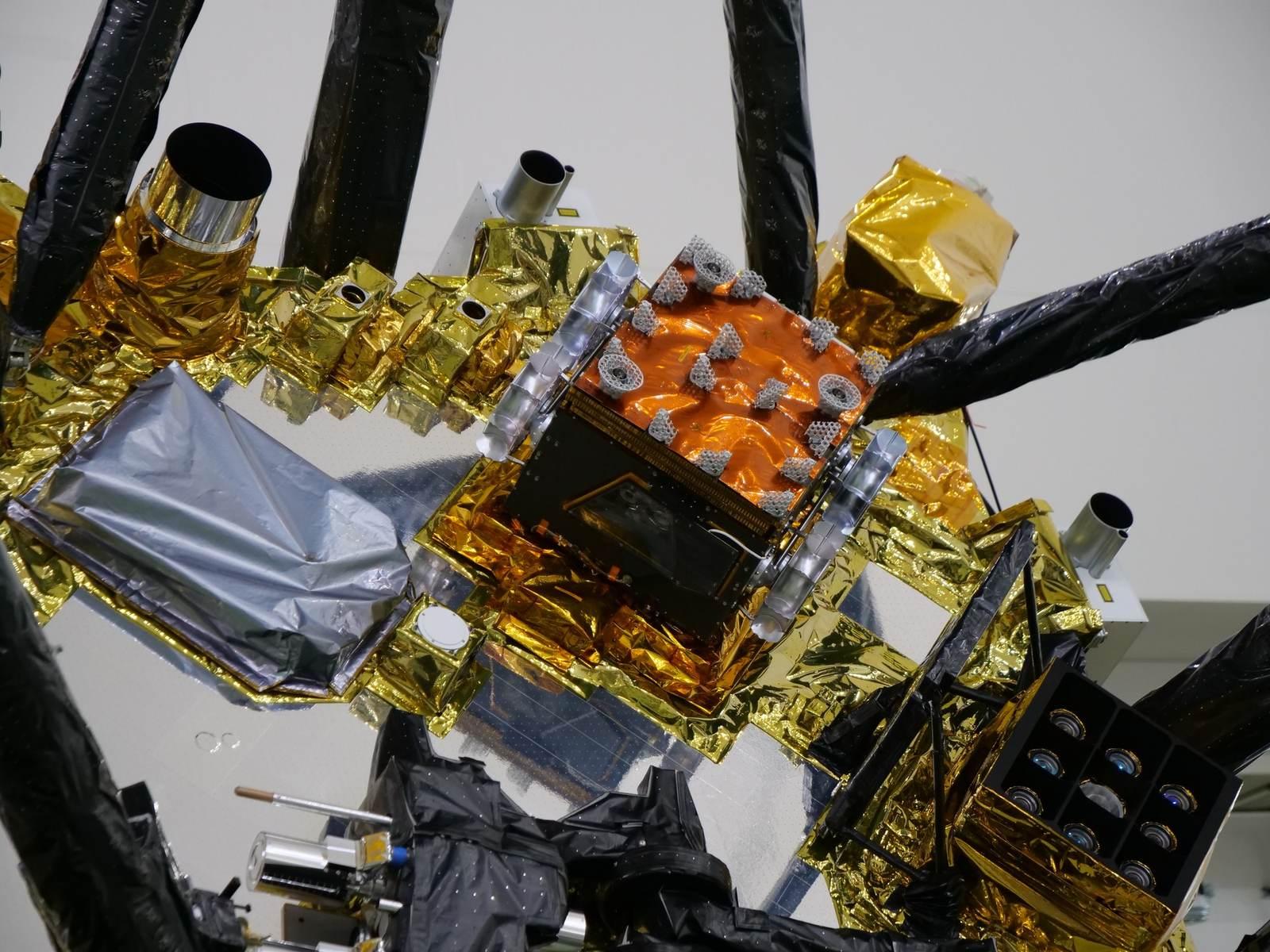 The IDEFIX rover fitted between the landing legs of the MMX exploration module in a cleanroom, with a person in a lab coat pointing at its location.
