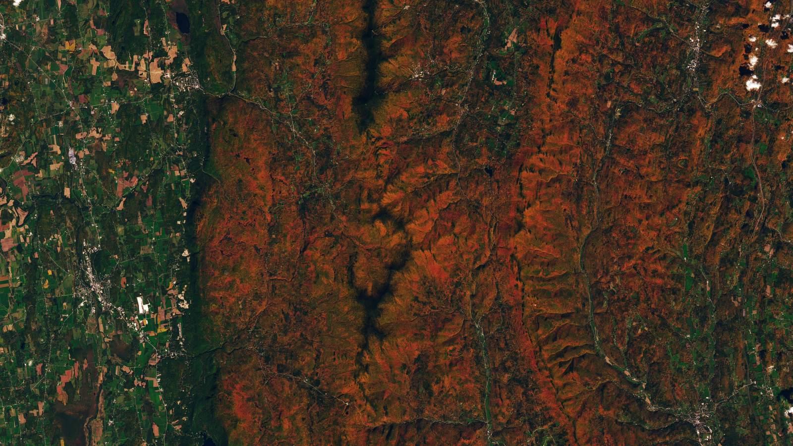 Satellite view of forested ridges in central Vermont glowing red and orange, with the river valleys and lower ground still green.