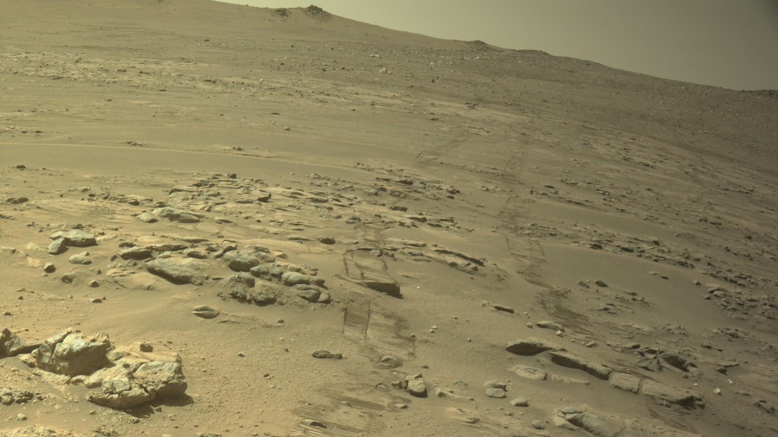 Vue d'une caméra de navigation du rover martien : au premier plan le pont du rover et ses instruments, au-delà un sol rocheux ocre et gris qui descend vers une plaine poussiéreuse et des reliefs lointains.