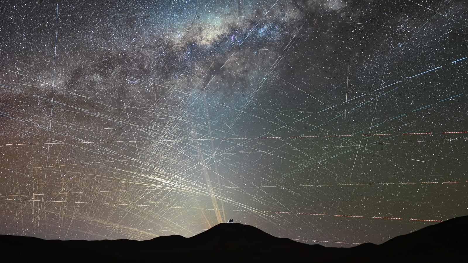 A starry night sky crossed by dozens of long, parallel and slanted streaks, above the dark silhouette of a mountain desert and a small metallic dome.