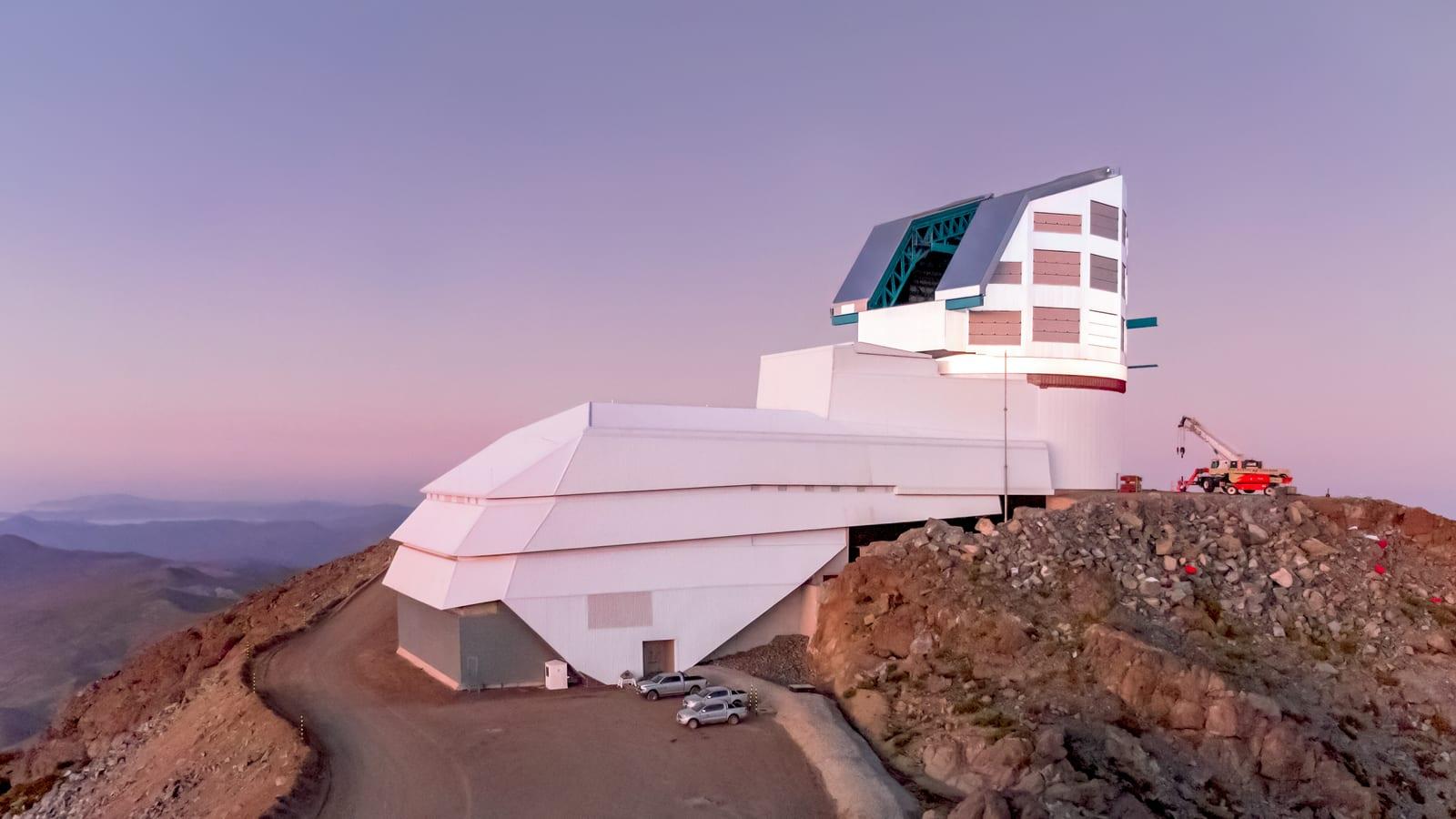 A dawn view of the Vera C. Rubin Observatory, a closed metallic dome on a rocky ridge under a sky fading from blue to orange.