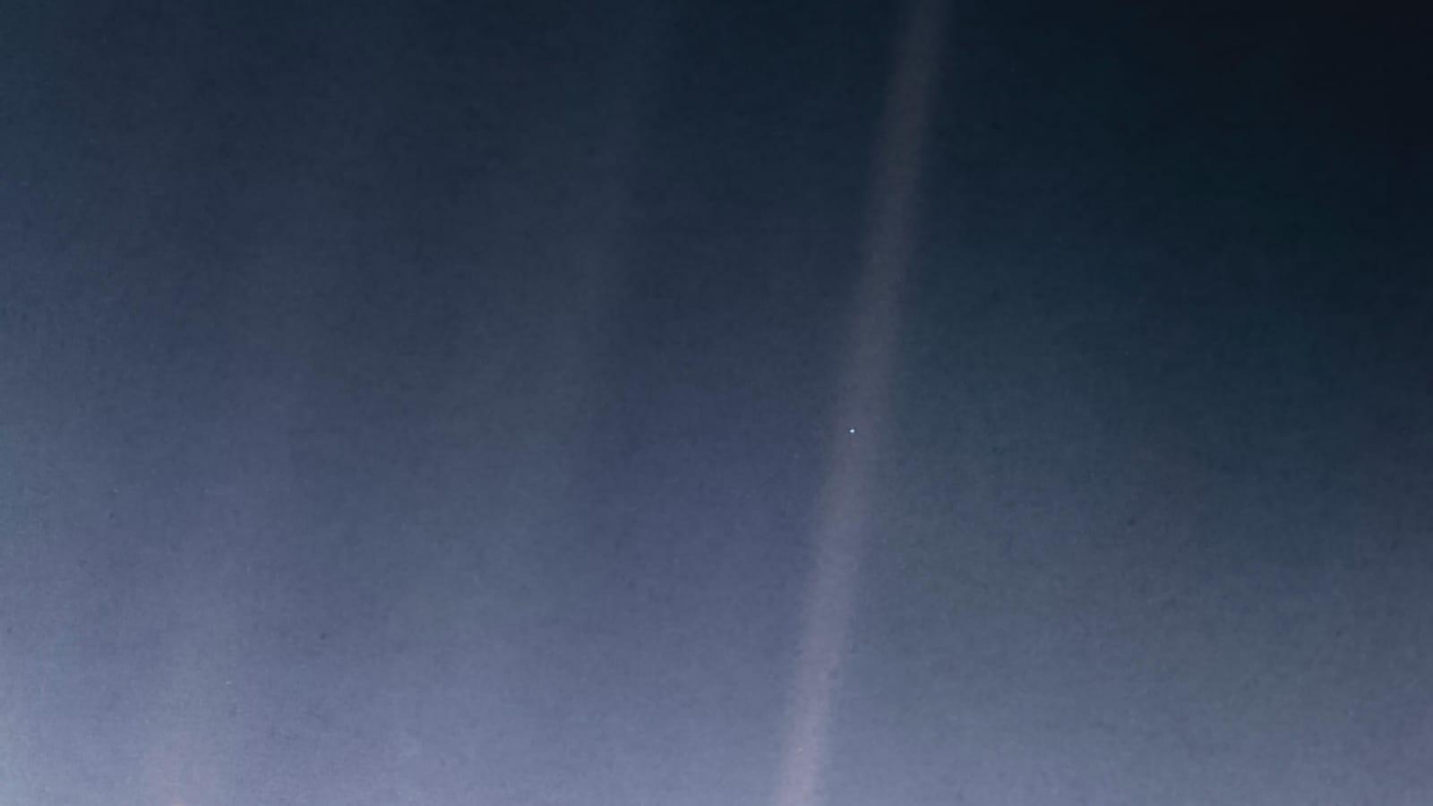 Image reprocessed by NASA in 2020: a narrow diagonal beam of light cuts across black space, with a single pale blue dot at its center, Earth photographed from 3.7 billion miles away.